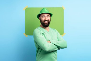 Yeşil şapkalı ve gömlekli sakallı adam Aziz Patrick Günü 'nü kutluyor. Kolları mavi bir duvarda yeşil bir zemine bağlı.