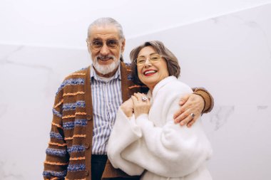 Yaşlı çift sıcak bir kucaklaşmayı paylaşıyor, her ikisi de ışıl ışıl gülümsüyor. Neşeli ifadeleri, uzun süreli ilişkilerin güzelliğini vurgulayarak mutluluğu ve arkadaşlığı ifade eder.