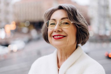 Kısa gri saçlı ve gözlüklü zarif son sınıf öğrencisi şehir ortamında güvenle gülümsüyor. Beyaz bir önlük giyerek hareketli bir şehir zemininde sıcaklık ve pozitiflik yayıyor. Moda konsepti