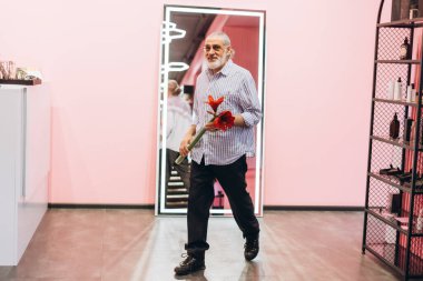Beyaz sakallı yaşlı bir adam modern, şık bir iç mekanda yürürken neşeyle çiçekleri tutuyor. Ayar büyük bir ayna ve yumuşak pembe duvarlar içeriyor, sıcak bir atmosfer yaratıyor.