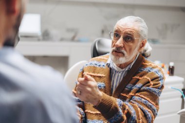 Yaşlı bir adam, modern bir klinikte bir doktorla tıbbi konsültasyon sırasında yaşlı yetişkinler için etkili iletişim ve sağlık hizmetlerine odaklanarak belirtileri anlatırken el kol hareketi yapıyor.