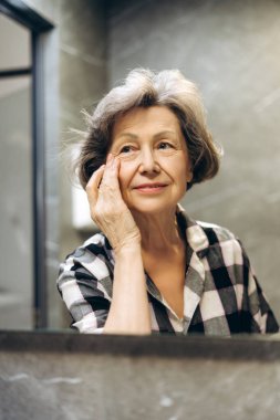 Modern banyo aynasına yansıyan zarif, olgun bir kadın nazikçe yüzüne dokunuyor. Kendine güveni ve zarafeti var, sakin bir ortamda kareli gömlek giyiyor. Cilt bakımı kavramı