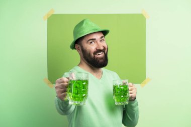 Bearded man with green hat and sweater holding two mugs of green beer celebrating St. Patrick's Day on green background with copy space clipart