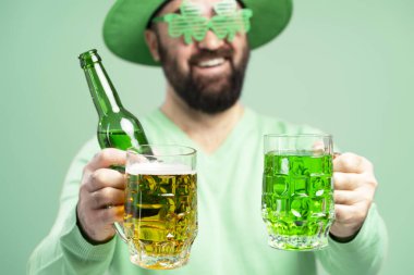 Yeşil Aziz Patrick Günü gözlüğü ve şapkası takan sakallı adam elinde yeşil arka planda yeşil bir şişe bira ve bir bardak yeşil bira tutuyor.
