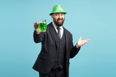 Bearded businessman wearing suit and green hat toasting with green beer on light blue background for St. Patrick's Day clipart