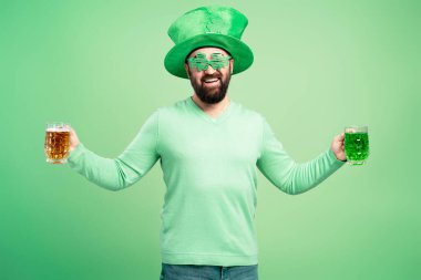 Aziz Patrick Günü şapkası ve gözlüğü takan mutlu bir adam elinde bir bardak bira ve bir bardak yeşil bira ile Aziz Patrick Günü 'nü kutluyor.