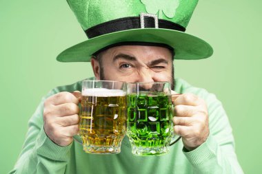 Sakallı adam iki bira bardağı tutuyor, biri yeşil biralı yeşil leprikon şapkalı yeşil arka planda