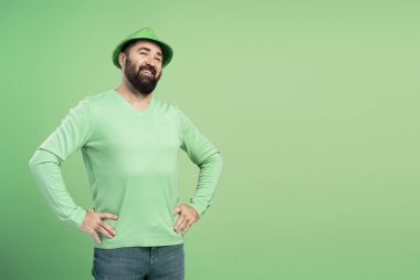 Mutlu sakallı adam yeşil giyinerek Aziz Patrick Günü 'nü kutluyor. Elleri kalçasında, gülümsüyor ve yeşil bir arka plana bakıyor.