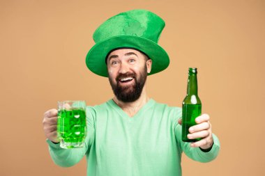 Happy man with beard wearing green leprechaun hat offering bottle and glass of green beer celebrating St. Patrick's Day clipart