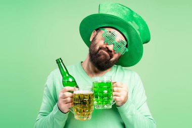 Sakallı, cüce şapkalı ve yonca şeklinde gözlüklü bir adam elinde bir kupa yeşil bira ve Aziz Patrick gününü kutlamak için bir şişe bira tutuyor.