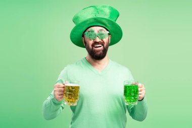 Aziz Patrick Günü 'nü kutlayan mutlu adam elinde normal bir bira ve yeşil bir bira yeşil arka planda komik bir şapka ve yonca şeklinde gözlük takıyor.