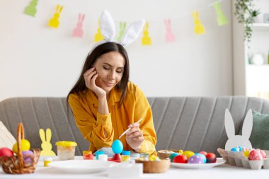 Paskalya yumurtalarını dikkatlice çizen kadın tavşan kulaklığı takıyor, Paskalya tatilinin tadını çıkarıyor.