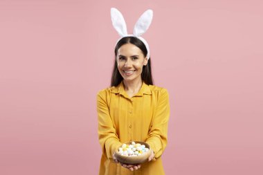 Tavşan kulaklı, gülümseyen bir kadının portresi. Pembe arka planda renkli paskalya yumurtaları sunuyor.