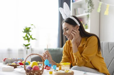 Kadın evde paskalya yumurtalarını dikkatlice boyuyor, tavşan kulağı takıyor ve gülümsüyor, paskalyayı kutluyor.