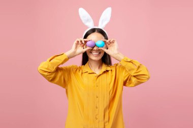 Paskalya yumurtalarıyla gözlerini kapayan, tavşan kulaklığı takan ve Paskalya bayramını kutlayan gülümseyen kadının stüdyo portresi.