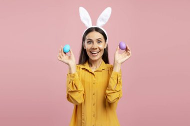 Tavşan kulaklı heyecanlı kadın pembe arka planda iki renkli Paskalya yumurtası tutuyor.
