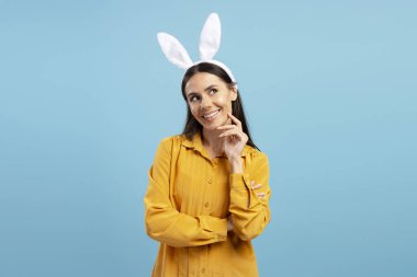 Paskalya tavşanı kulaklı, turuncu gömlekli düşünceli bir kadın mavi arka plan üzerinde kafa yoruyor.