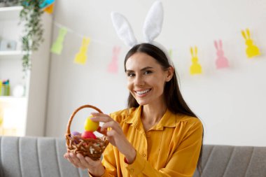 Tavşan kulaklığı takan gülümseyen kadın renkli paskalya yumurtaları içeren küçük bir hasır sepet tutuyor.
