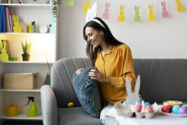 Tavşan kulağı takan gülümseyen kadın kanepede oturuyor ve süslü oturma odasında yastığın altına gizlenmiş paskalya yumurtaları buluyor.