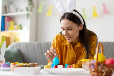 Paskalya yumurtalarını dikkatlice çizen kadın, arka planda paskalya süsleri asılı.