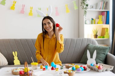 Kanepede oturan, tavşan kulağı takan ve evde paskalya yumurtası çizen bir kadın. Paskalya tatilinin tadını çıkarıyor.