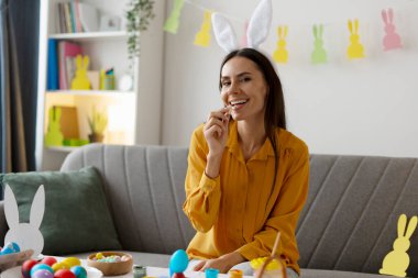 Evde Paskalya kutlaması için yumurta boyayıp dekore ederken çikolata yiyen tavşan kulaklı genç bir kadın.
