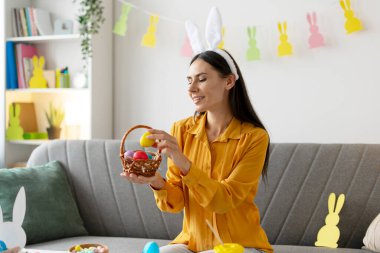 Evde Paskalya kutlaması sırasında elinde renkli yumurtalarla sepet tutan tavşan kulağı takan gülümseyen bir kadın.