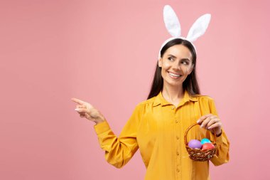 Mutlu kadın parmak göstererek fotokopi alanını işaret ediyor tavşan kulaklığı takıyor ve pembe arka planda renkli yumurtalarla dolu bir Paskalya sepeti tutuyor.