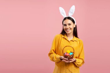 Tavşan kulaklığı takmış, gülümseyen bir kadının portresi pembe arka planda paskalya yumurtası sepeti tutuyor.