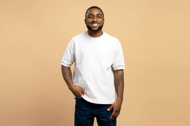 Elleri cebinde gülümseyen, bej arka planda beyaz tişört giyen Afro-Amerikan bir adamın stüdyo portresi.