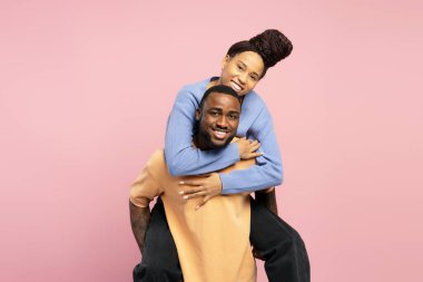 Genç siyahi kadın erkek arkadaşını kucaklıyor, pembe stüdyo geçmişinde gezintiden keyif alıyor, mutlu bir milenyum çifti birlikte eğleniyor.