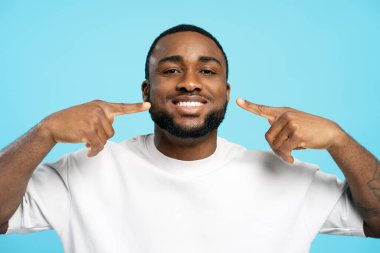 Genç adam pırıl pırıl gülümserken mükemmel beyaz dişlerini parlak mavi bir arka planda göstererek kendine güven ve neşe saçıyor.