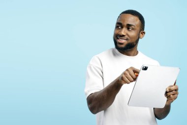 Genç Afrikalı Amerikalı adam dijital tablet kullanarak ekranı işaret ediyor. Mavi arka planda gülümsüyor.