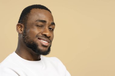 Neşeli, Afro-Amerikan bir adamın bej bir arka planda kameraya göz kırpan portresi pozitifliği ve arkadaşlığı ifade ediyor.