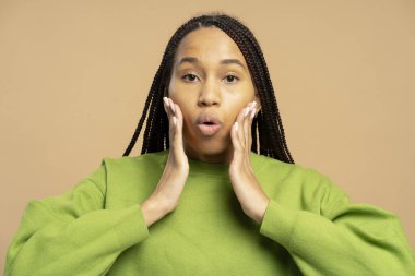 Uzun örgülü, yeşil kazaklı, yanaklarını elleriyle tutup bej bir arka planda ağzını açan genç bir kadının stüdyo portresi.