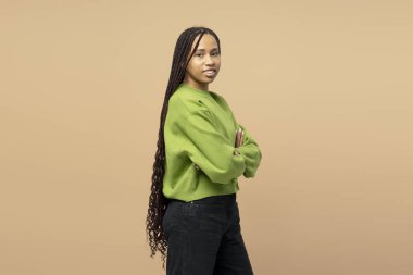 Uzun örgülü saçlı, yeşil süveterli, bej arka planda kolları çapraz duran genç bir kadının stüdyo portresi.