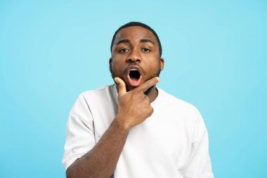 Beyaz tişörtlü Afro-Amerikalı genç adam çok şaşırdı. Mavi arka planda parmağını açık ağzına doğrultuyor.