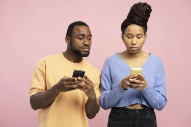 Akıllı telefon kullanan genç bir çiftin stüdyo fotoğrafı. Adam kadının telefonuna tuhaf bir ifadeyle bakıyor.