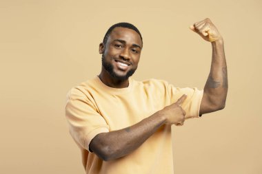 Genç adam kaslarını esnetirken geniş bir gülümseme ve gururla kaslı kolunu işaret ederek kendine güven ve güç yayıyor.