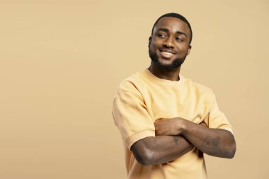 Kollarını kavuşturup gülümseyen, bej rengi bir arka planda kendine güvenini ve pozitifliğini ifade eden Afrikalı genç bir Amerikalının stüdyo portresi.