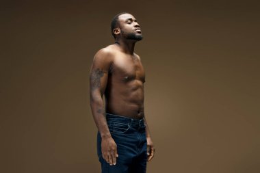 Dövmeleri olan, gözleri kapalı, kot giyen ve çıplak gövdesini gösteren siyahi bir adamın stüdyo fotoğrafı.