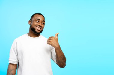 Neşeli afro-amerikan adam başparmağını kaldırıyor ve mavi stüdyonun arka planında işaret ediyor.