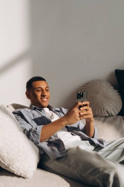 Mutlu Latin adam cep telefonunu evde rahat bir kanepeye uzanıp internet iletişiminin, internetin, alışverişin ya da video izlemenin tadını çıkarırken kullanıyor. Çevrimiçi teknoloji kavramı