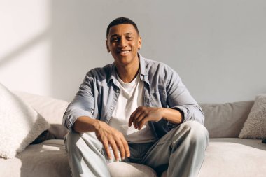 Evdeki rahat kanepede otururken gülümseyen genç, gülümseyen Afrikalı Amerikalı adam. Moda modeli kameraya bakıyor. Reklam kavramı
