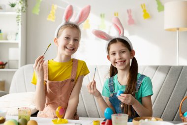 Tavşan kulaklığı takan iki gülümseyen kız masaya oturmuş paskalya yumurtası çiziyor.