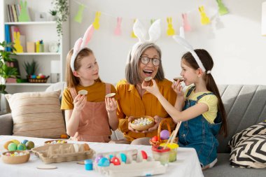 İki mutlu ikiz kardeş tavşan kulağı takıyor büyükanneleriyle çikolatalı yumurtalarını paylaşıyor, evdeki kanepede oturuyor.