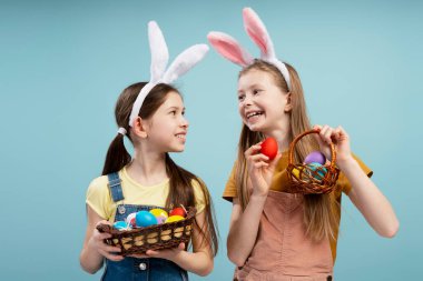 Tavşan kulağı takan iki mutlu genç kız ellerinde renkli Paskalya yumurtalarıyla dolu hasır sepetler ve açık mavi bir arka planda birbirlerine gülümsüyorlar.