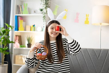 Kırmızı Paskalya yumurtasını gözüne doğru tutarken gülümseyen genç bir kadın, sepetteki renkli yumurtalarla çevrili, tavşan kulakları takıyor.