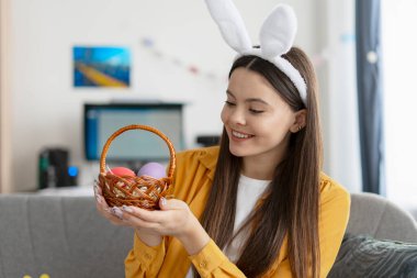 Tavşan kulaklığı takan genç bir kadın içinde paskalya yumurtaları olan küçük bir hasır sepet tutuyor ve gülümsüyor.