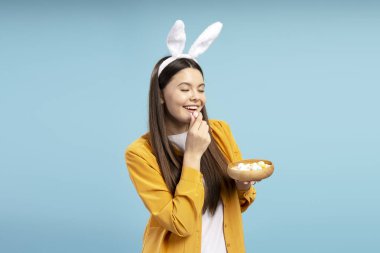 Tavşan kulaklı mutlu genç kadın mavi arka plandaki ahşap kaseden renkli paskalya yumurtaları yiyor.